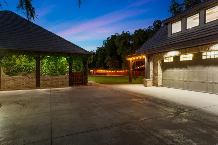 Expansive concrete driveway servicing a detached garage with stone accents, a dormer window feature, and a separate covered structure with brick detailing and lush greenery