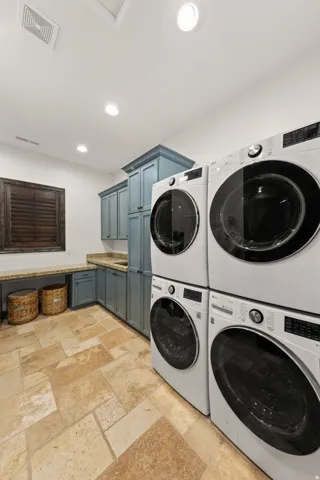 Laundry room featuring two sets of stacked front-load machines, light stone tile flooring, and blue-painted cabinetry