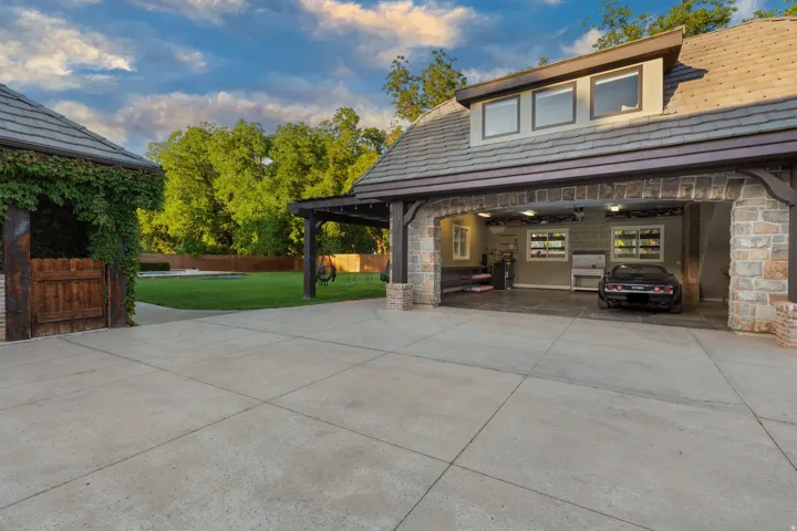 Stone and wood-beam carport with a second-story dormer, featuring a concrete driveway, a manicured lawn, and a secondary structure with a wooden gate and vine-covered pergola