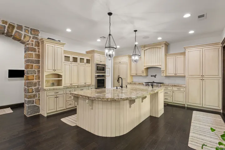 Gourmet kitchen featuring a large curved island with a stone-finish countertop, cream-toned cabinetry, dark wood-finish flooring, a stone archway, and stainless steel built-in appliances