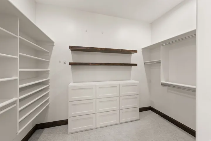 Spacious walk-in closet featuring extensive built-in shelving, two dark wood-finish wall shelves, a white built-in nine-drawer dresser, and ample hanging rod space