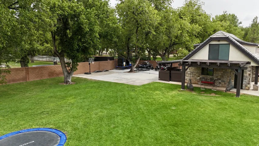 Expansive green lawn, mature trees, concrete sport court with basketball hoop, stone-clad exterior with pergola, and a brick perimeter wall