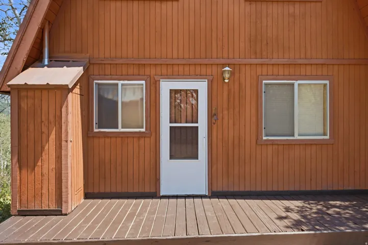 Exterior featuring vertical wood siding, a white entry door with screen, two window units, and a porch light fixture