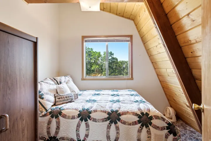 Bright bedroom featuring a large window, wood-finish flooring, wood plank accent wall, built-in closet with dark wood-finish door, and exposed ceiling beam