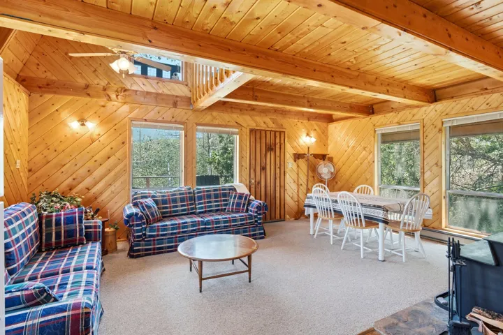 Wood-paneled living area featuring exposed beam ceilings, a central ceiling fan, multiple windows providing natural light, and a fireplace with a stone hearth