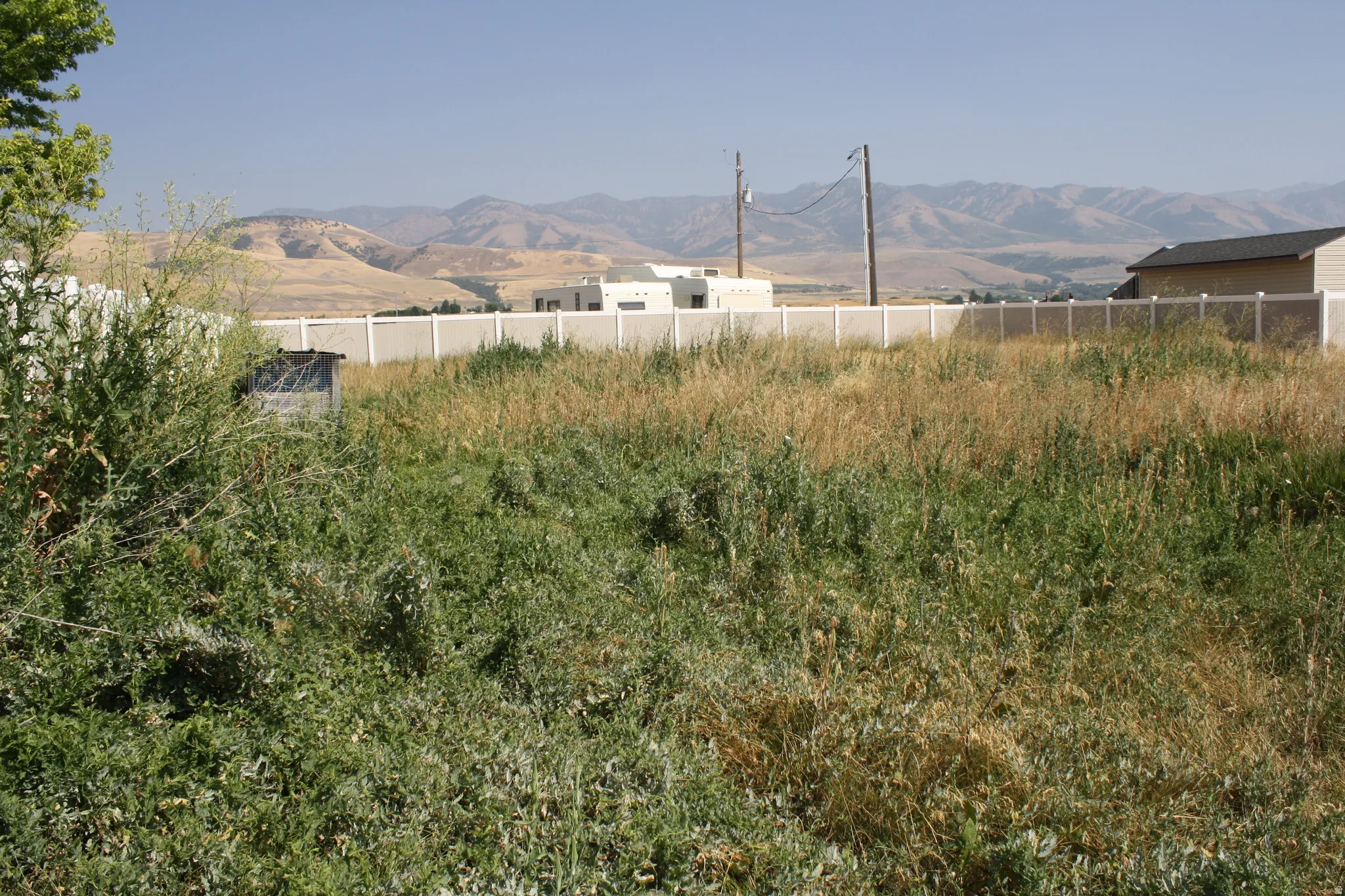 Expansive undeveloped lot featuring varied natural vegetation, a white privacy fence along the perimeter, and distant mountainous terrain