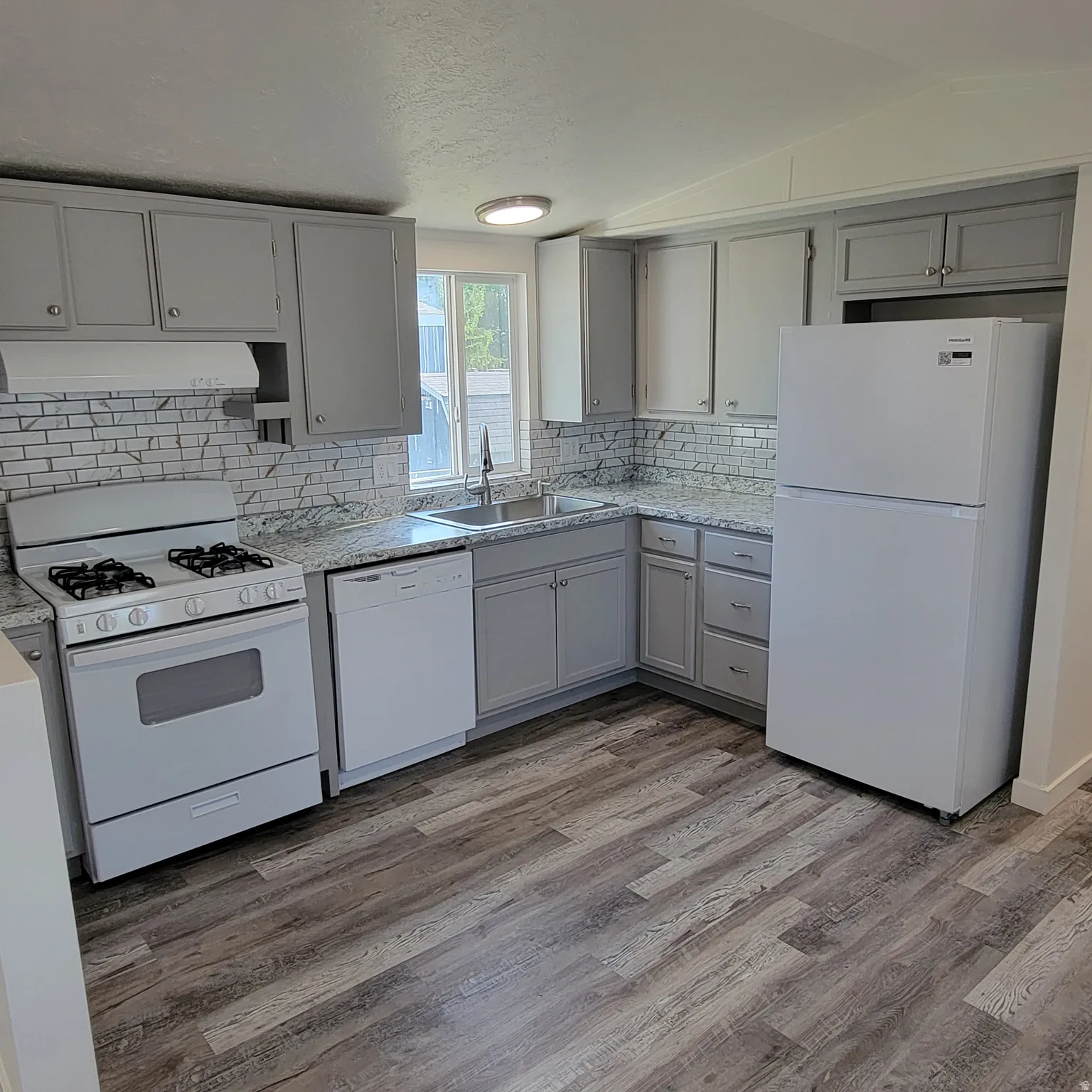 Kitchen featuring grey cabinetry, white appliance package, wood-finish flooring, subway tile backsplash, and a corner sink with window