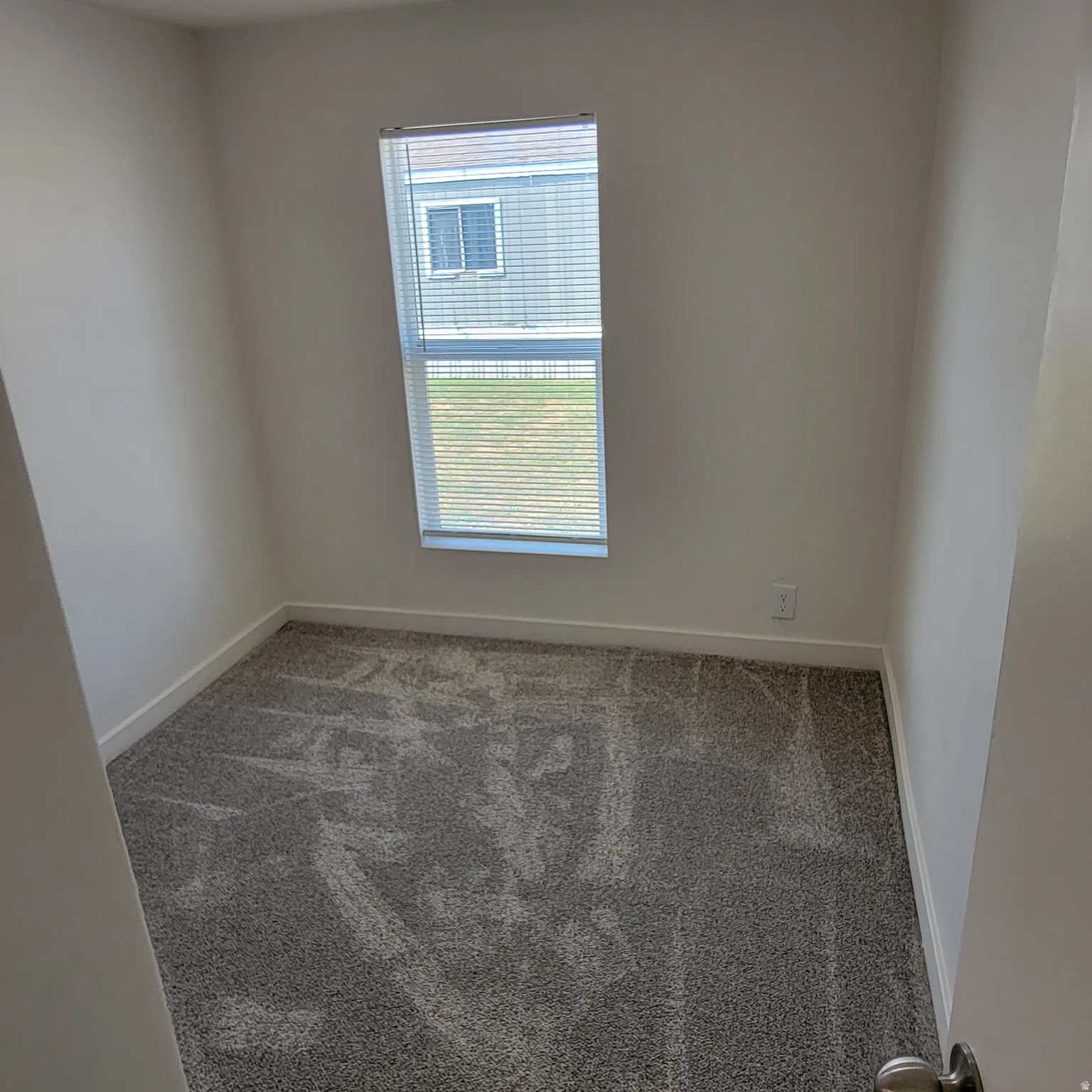 Neutral wall paint, gray patterned carpet, white baseboards, and a single window with horizontal blinds