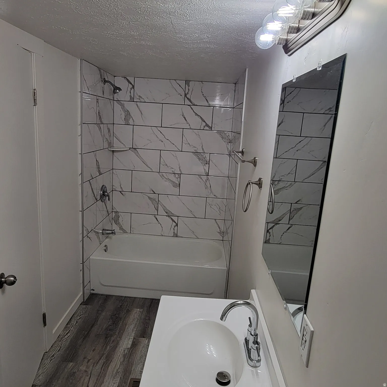Bathroom featuring a white bathtub with marble-look subway tile surround, chrome fixtures, wood-finish flooring, a white vanity with integrated sink, and a wall-mounted mirror