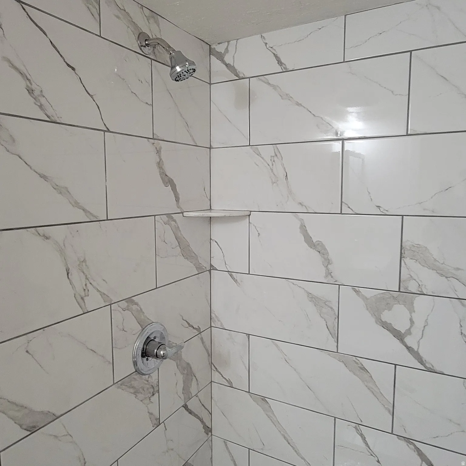 Shower enclosure featuring large format white tile with grey veining, chrome showerhead, chrome temperature control, and a built-in corner shelf