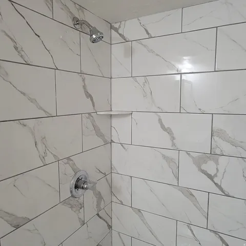 Shower enclosure featuring large format white tile with grey veining, chrome showerhead, chrome temperature control, and a built-in corner shelf