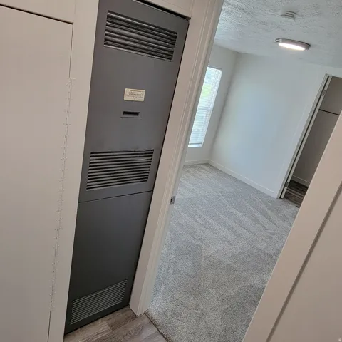 Interior utility closet with a dark gray louvered panel, light wood-finish flooring, and a bedroom beyond with light gray patterned carpeting, white walls, and a window with blinds