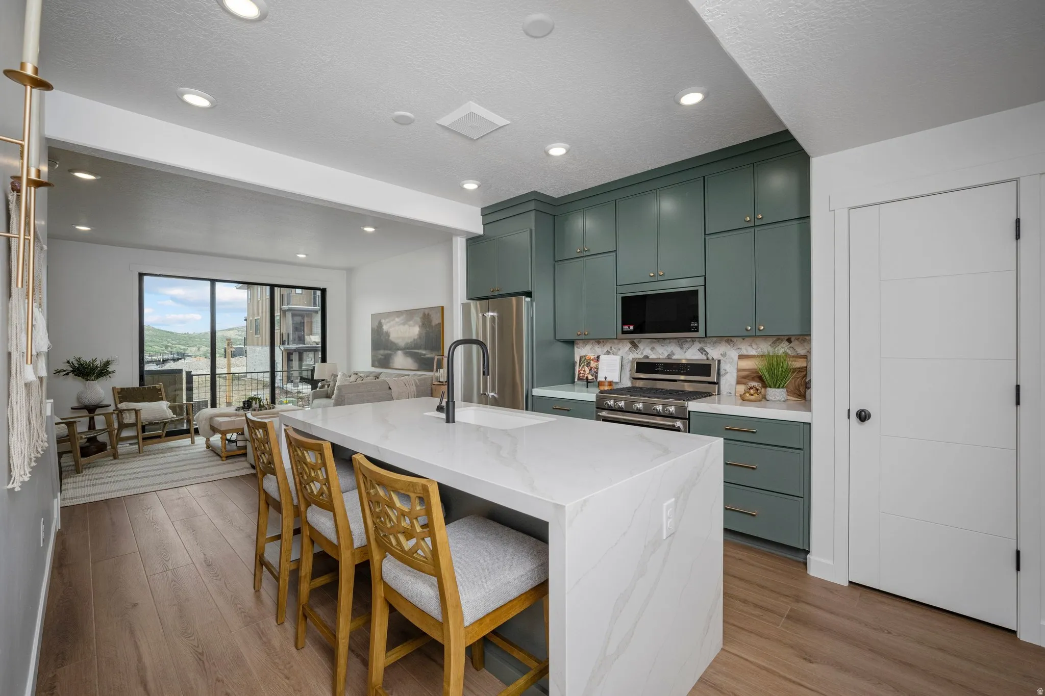 Modern kitchen featuring a large waterfall-edge island with seating, a stainless steel gas range, a mosaic tile backsplash, and contemporary green cabinetry with gold hardware