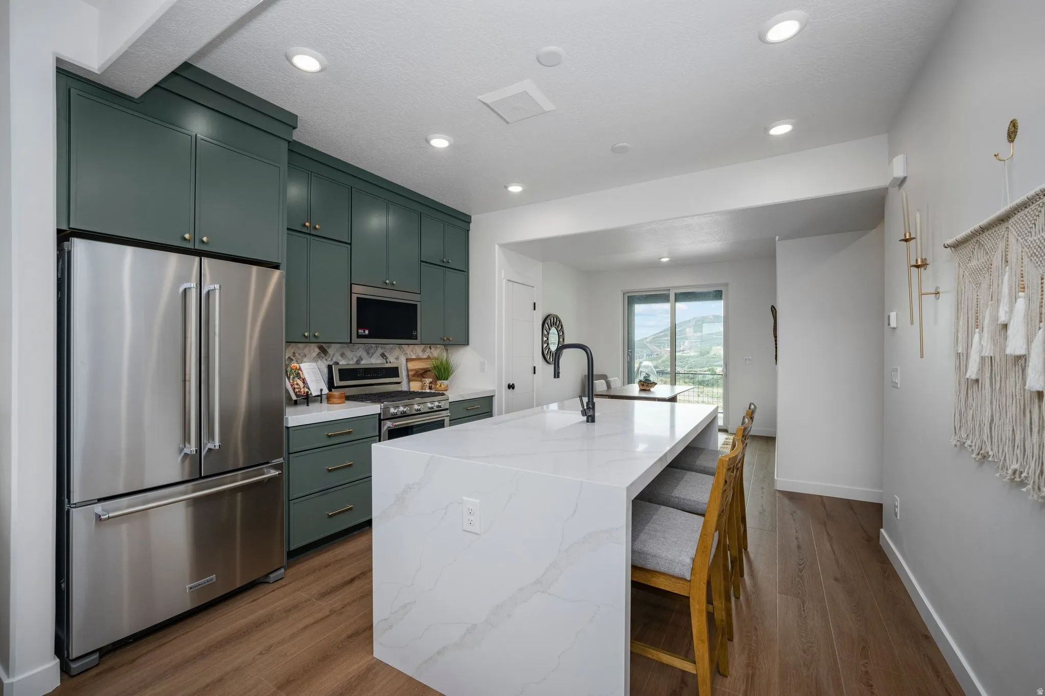 Modern kitchen featuring a waterfall island with integrated seating, stainless steel appliances, ample dark green cabinetry, a patterned backsplash, and wood-finish flooring