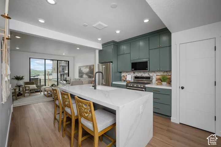 Modern kitchen featuring a large waterfall-edge island with seating, a stainless steel gas range, a mosaic tile backsplash, and contemporary green cabinetry with gold hardware