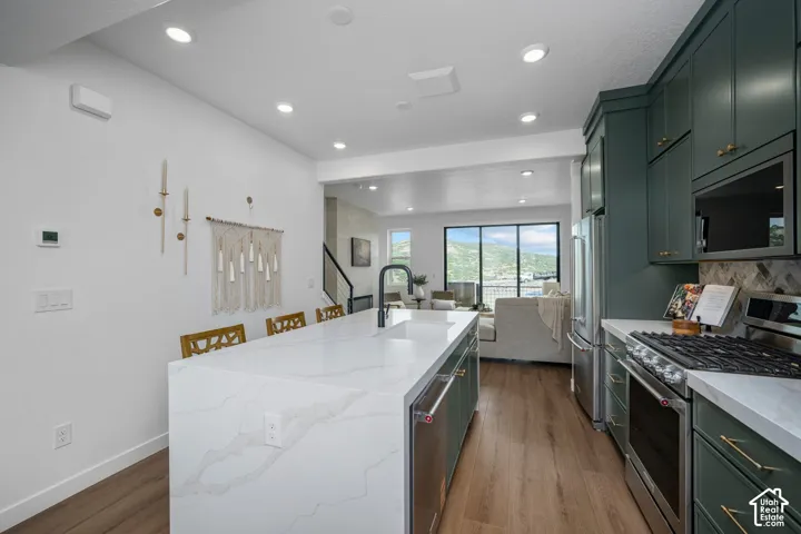 Kitchen featuring a waterfall island with an integrated sink, dark green cabinetry, stainless steel appliances, and wood-finish flooring