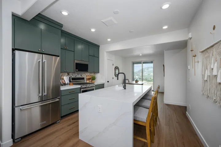Modern kitchen featuring a waterfall island with integrated seating, stainless steel appliances, ample dark green cabinetry, a patterned backsplash, and wood-finish flooring