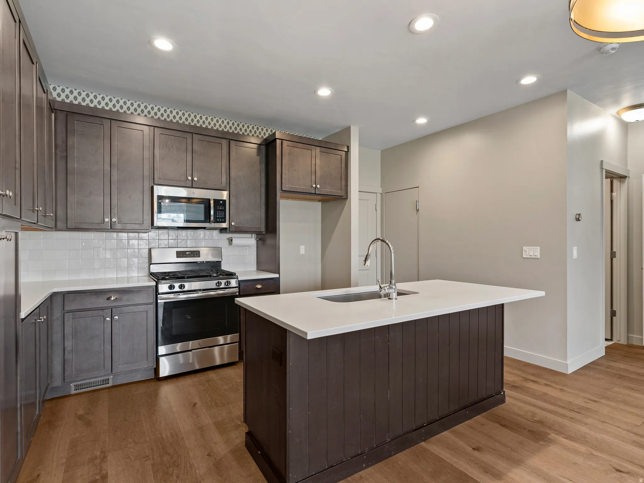 Kitchen featuring dark wood cabinetry, stainless steel appliances, a white tile backsplash, a central island with a sink, and wood-finish flooring