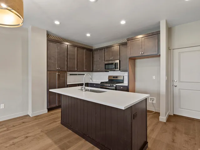 Modern kitchen featuring a central island with a solid surface countertop, dark wood cabinetry, stainless steel appliances, a white tile backsplash, and wood-finish flooring