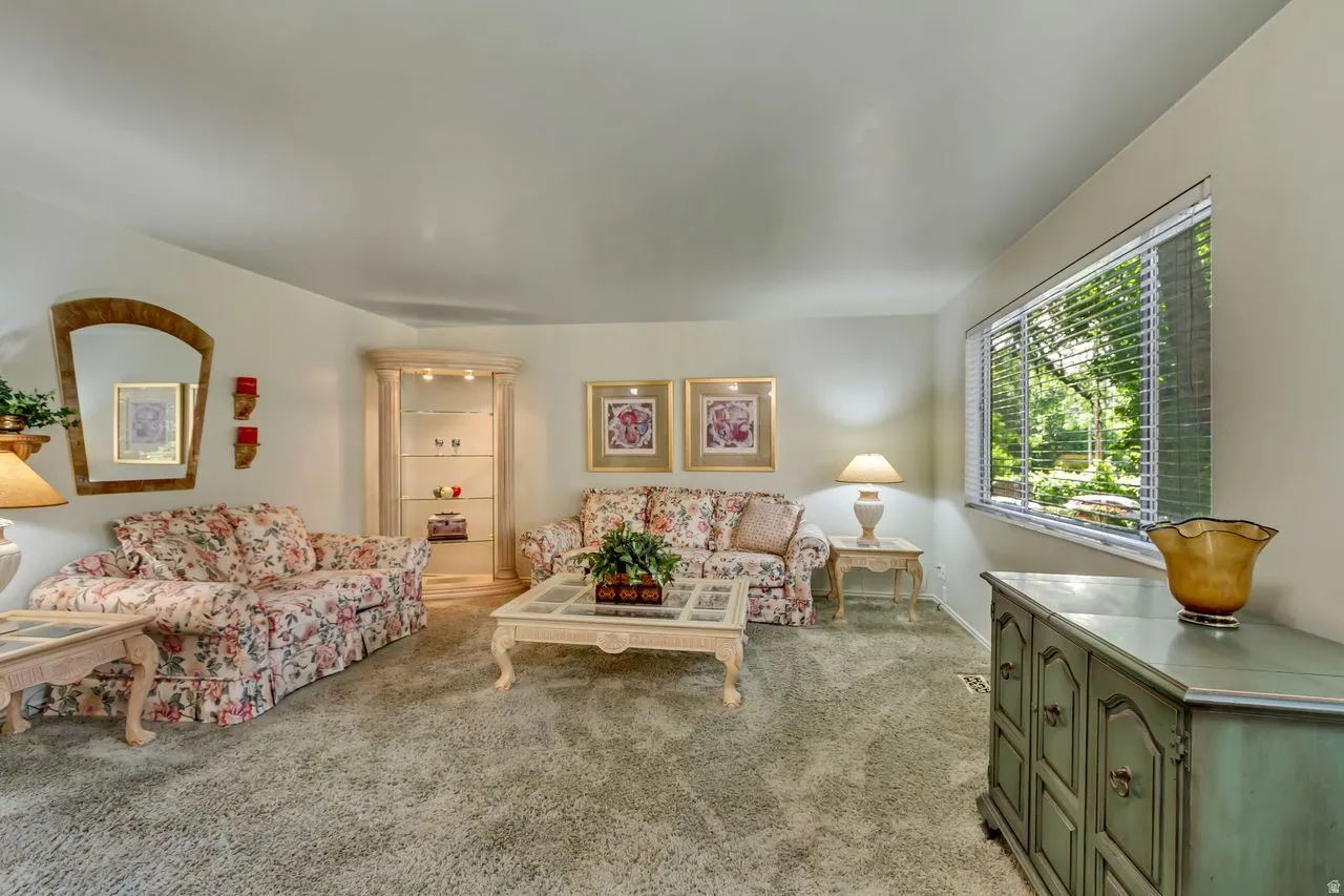 Spacious living area featuring light-toned walls, textured carpeting, and a large window with horizontal blinds