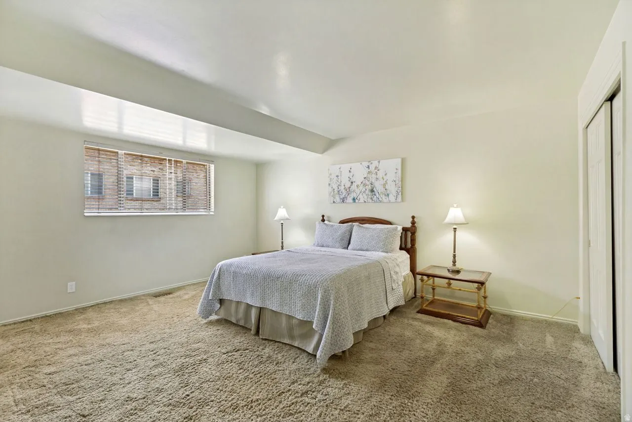 Carpeted interior room featuring a window with horizontal blinds, light-toned walls, and a white bi-fold closet door