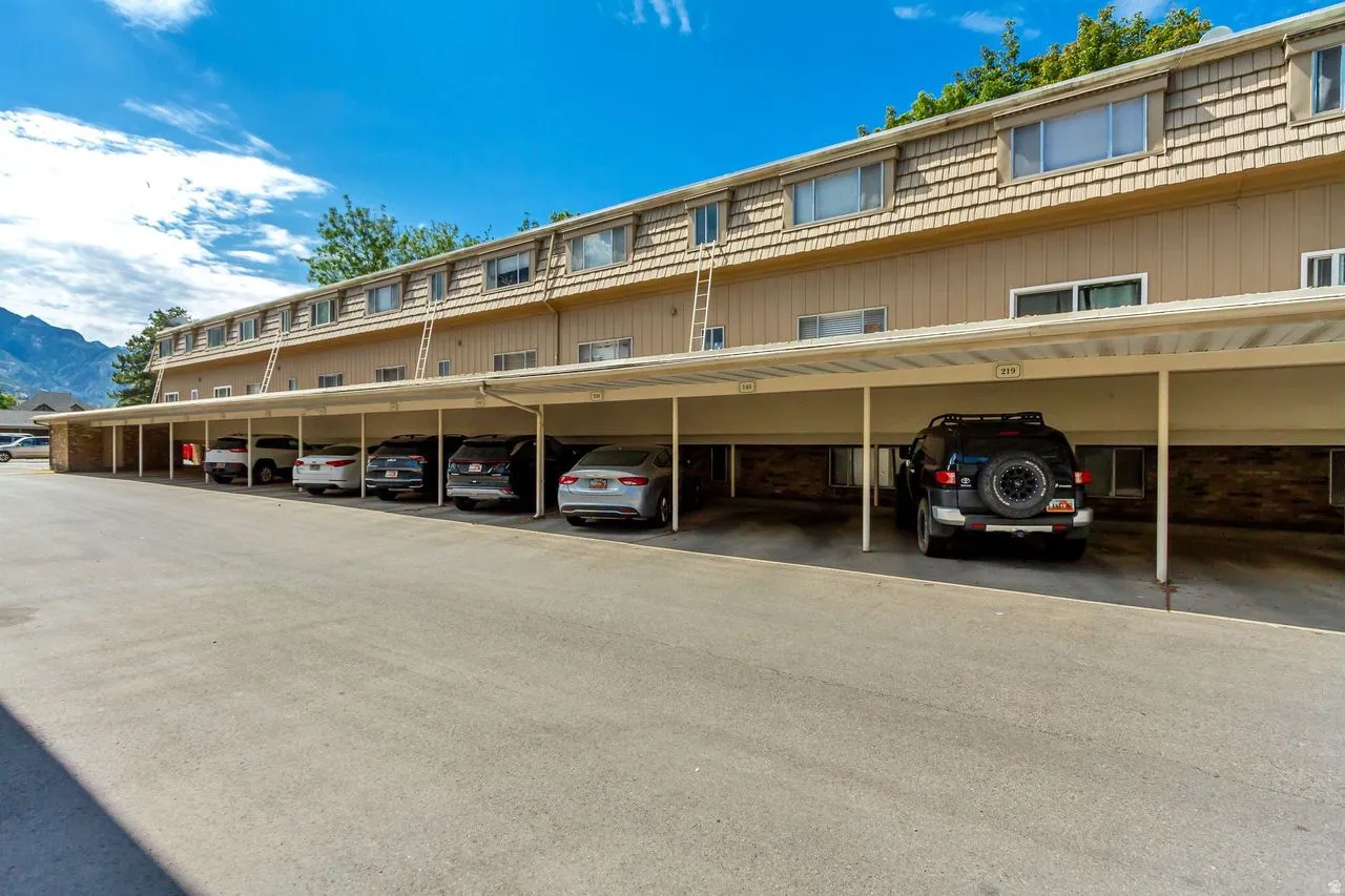 Covered parking structure with individual numbered spaces, featuring a paved lot and multi-story building facade with horizontal siding and shingle accents