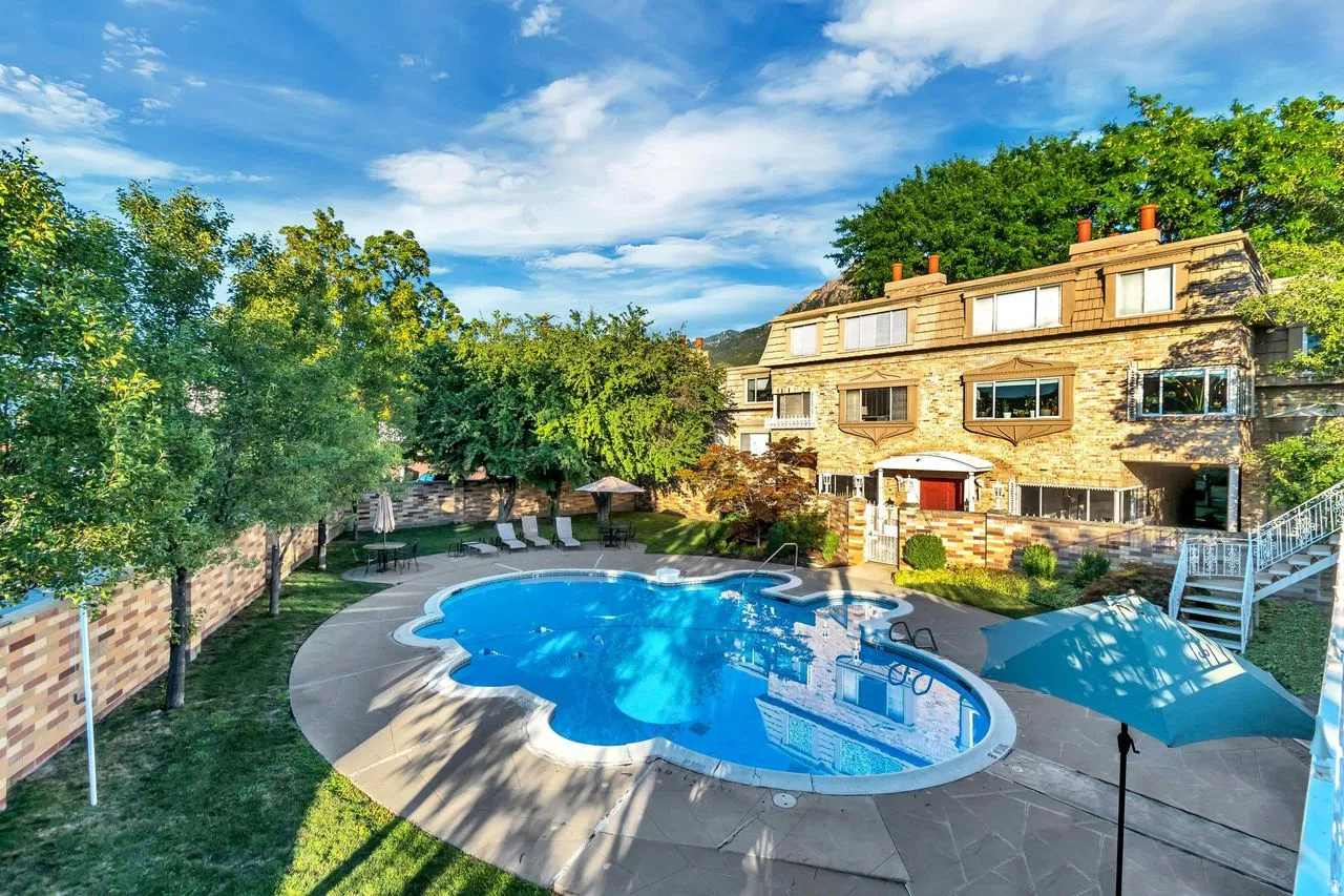 Resort-Style Backyard Oasis featuring a sparkling blue saltwater pool with integrated spa, curved sun deck with lounge seating areas, mature landscape trees providing natural shade, and a stunning stone-and-stucco residence with multi-level terraces overl