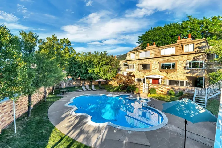 Resort-Style Backyard Oasis featuring a sparkling blue saltwater pool with integrated spa, curved sun deck with lounge seating areas, mature landscape trees providing natural shade, and a stunning stone-and-stucco residence with multi-level terraces overl