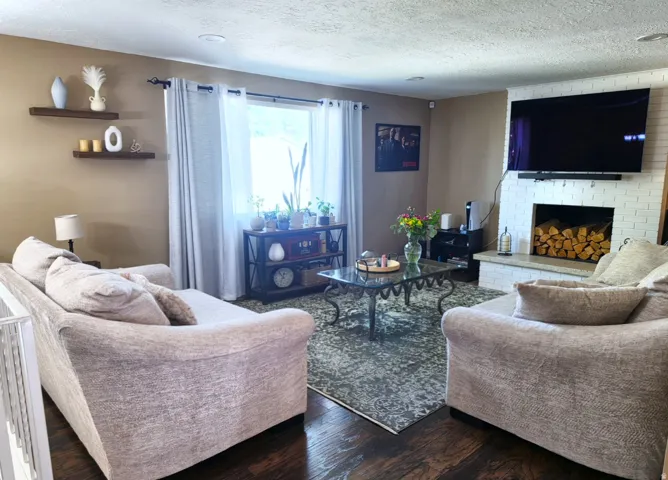 Living area featuring dark wood-finish flooring, a white painted brick fireplace with a stone hearth, and a large window with sheer drapes