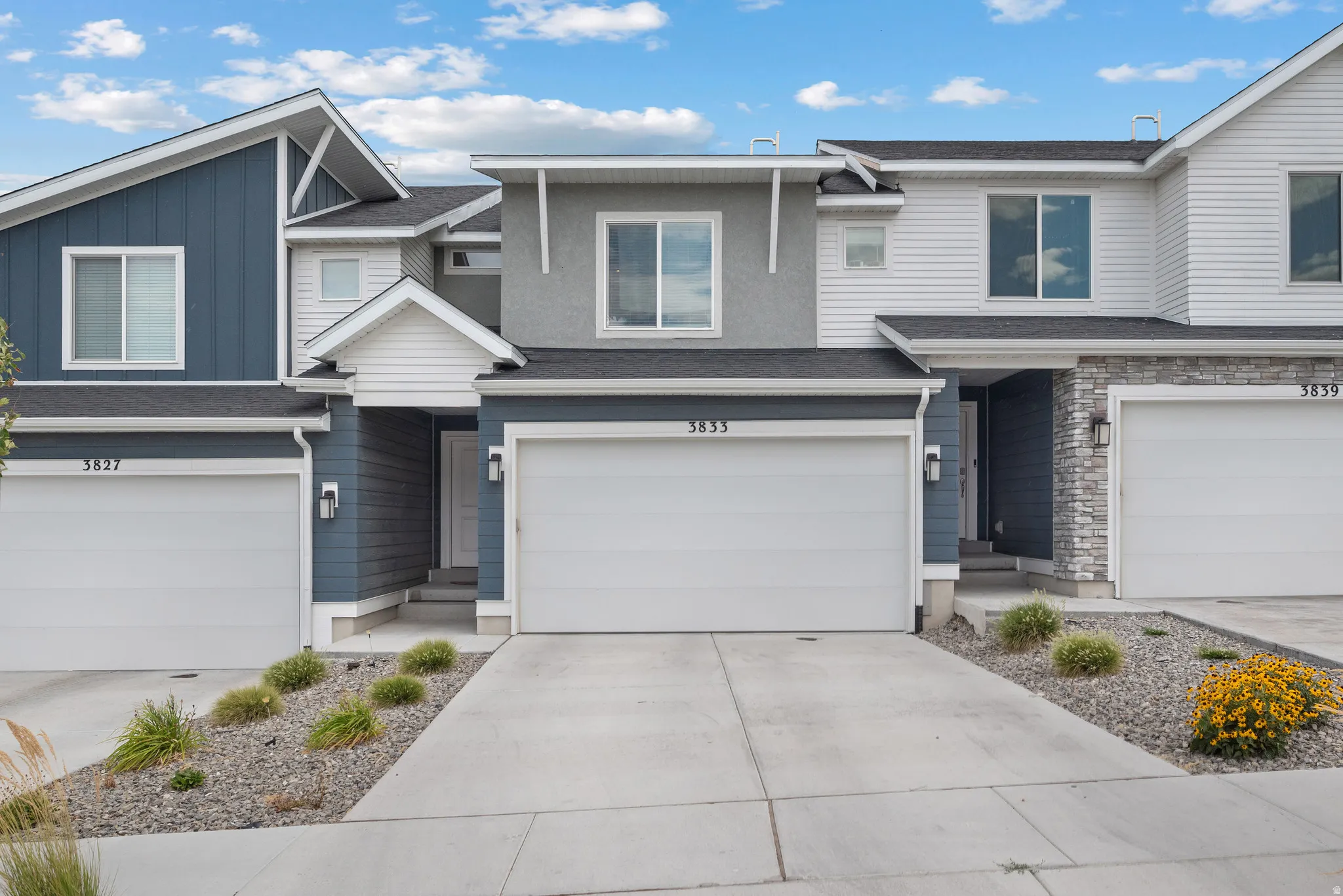 Modern townhome featuring a two-car garage with white paneled doors, contemporary exterior siding in neutral gray and white tones, pitched roofline with architectural detailing, stone accents on the facade, and a welcoming front entry with landscaped xeri