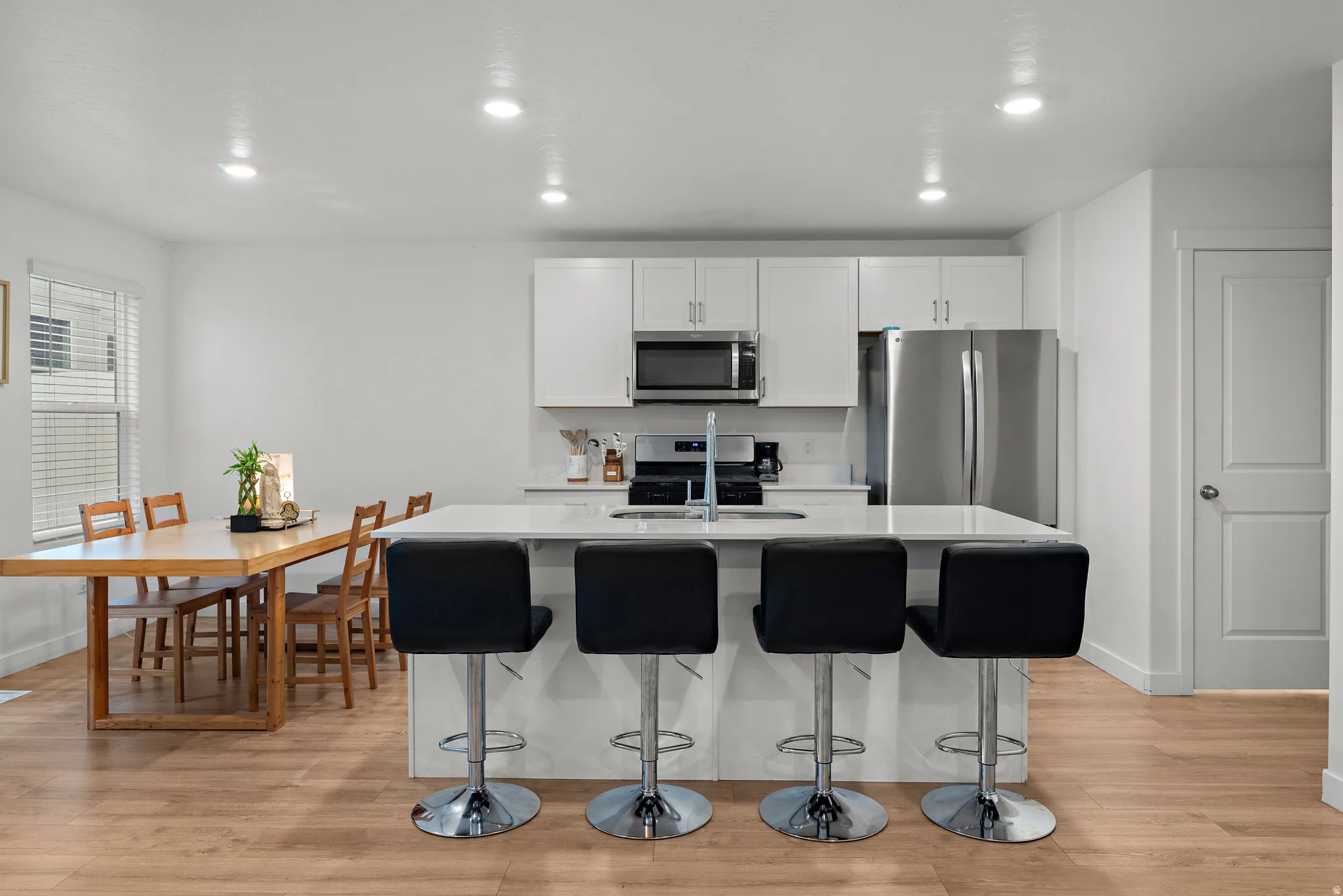 Modern open-concept kitchen and dining area featuring stainless steel appliances, white cabinetry with chrome hardware, a quartz countertop island with seating for four, recessed ceiling lighting, wood-finish flooring throughout, and a dedicated dining ta