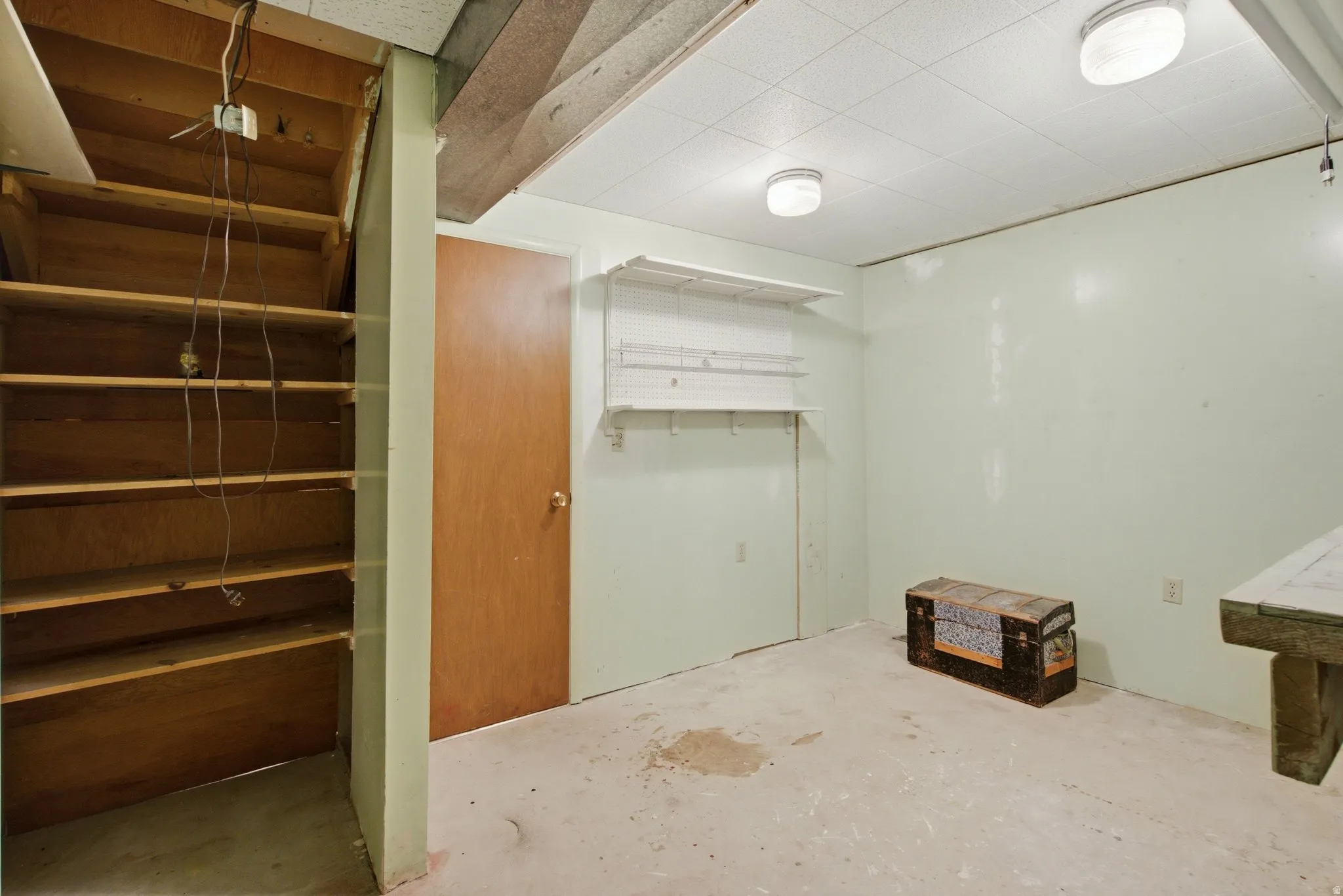 Unfinished basement space featuring a wooden staircase with integrated shelving, a solid core door, and exposed ceiling joists