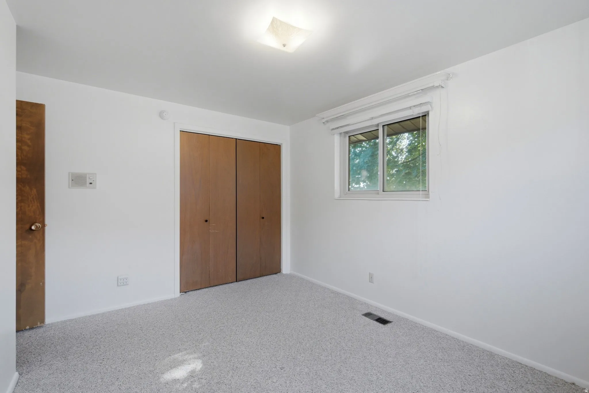 Bedroom with light gray carpeting, neutral white walls, and a window featuring a white-framed casement
