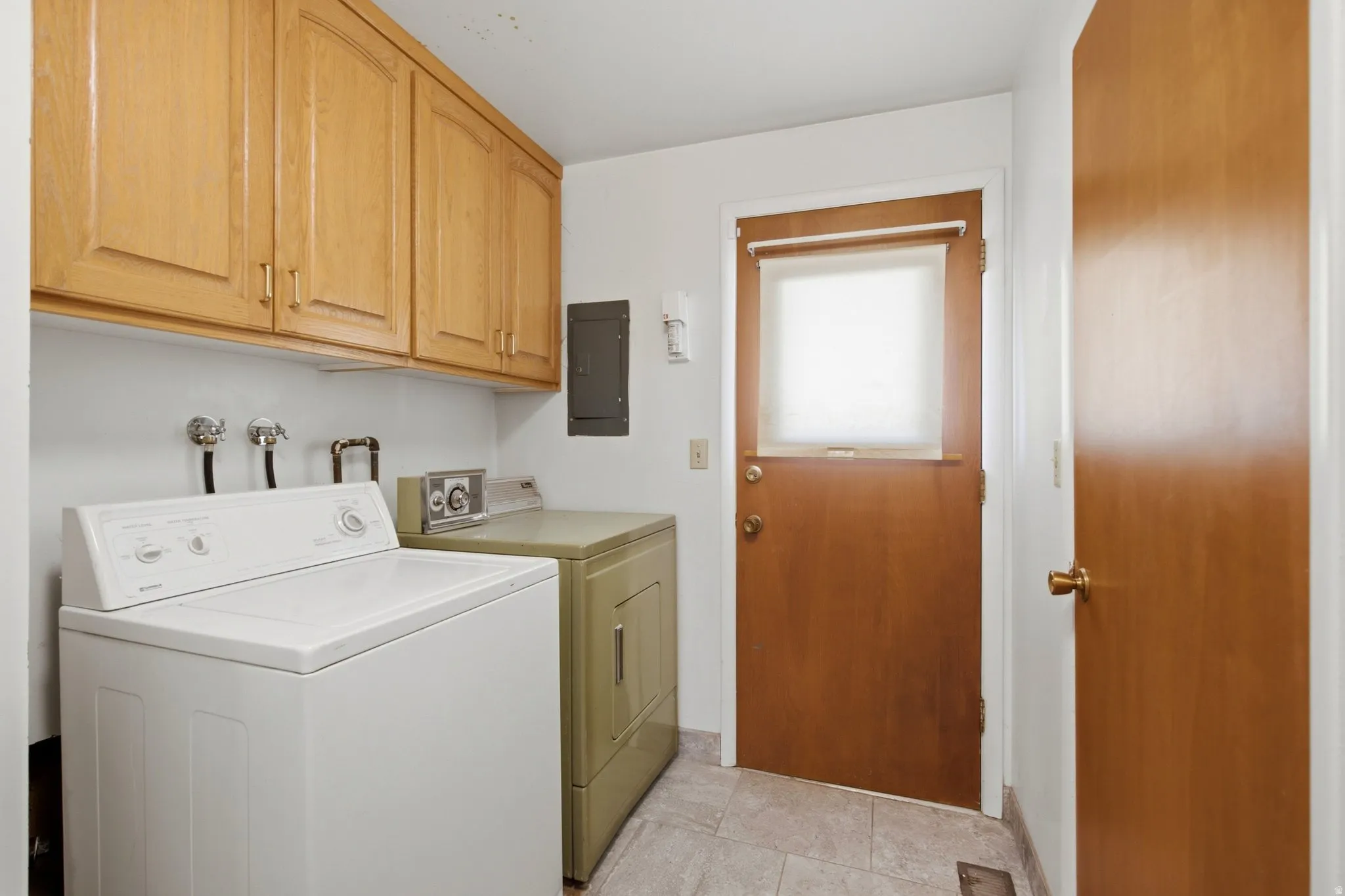 Dedicated laundry room featuring white front-load washer, olive-toned cabinetry with ample storage, tile flooring, and direct exterior access via wood-frame door with natural light from transom window