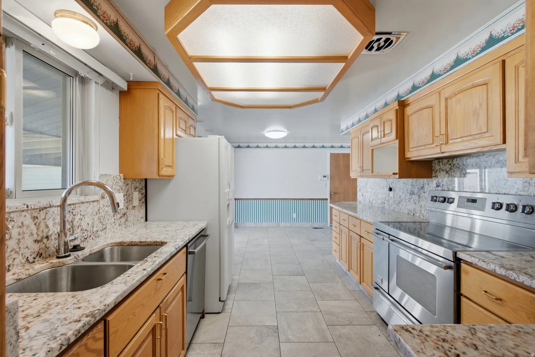 Expansive kitchen featuring light wood cabinetry, stone countertops, a double basin stainless steel sink with gooseneck faucet, tiled flooring, and a stainless steel electric range