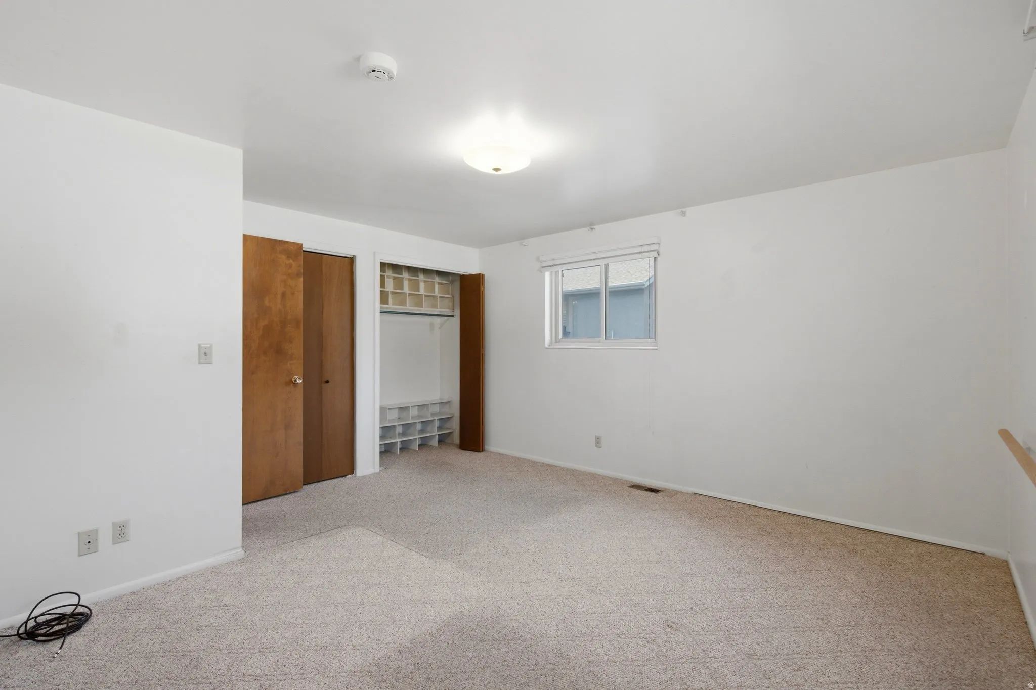 Spacious bedroom featuring neutral carpeting, two wood-finish closet doors with shelving, a large window with blinds, and multiple electrical outlets
