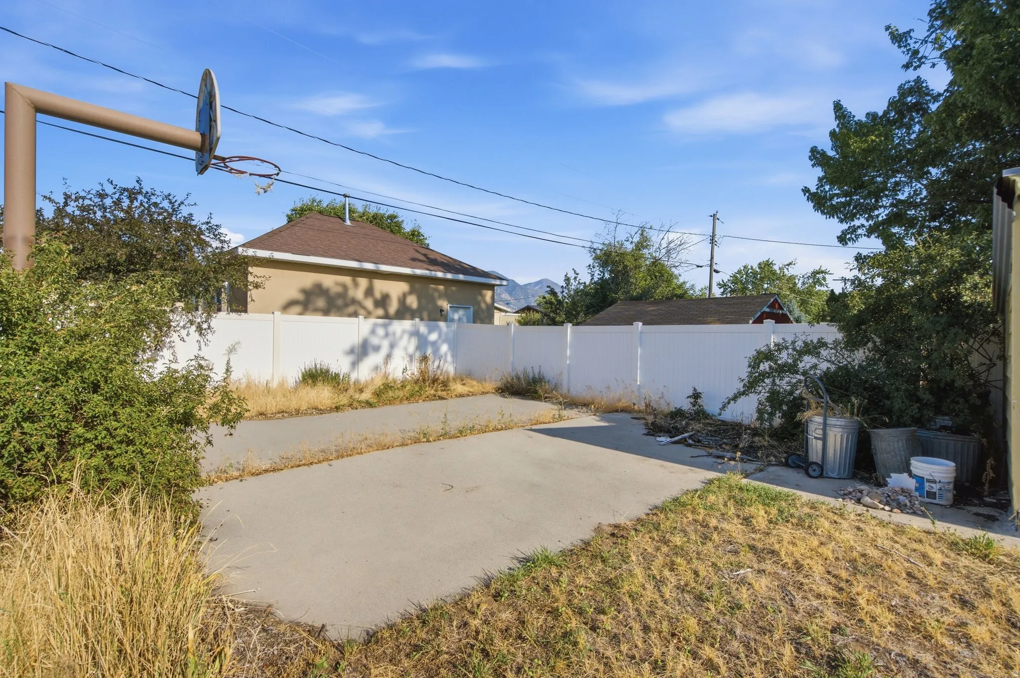 Concrete pad with an integrated basketball hoop, bordered by a white privacy fence and mature trees