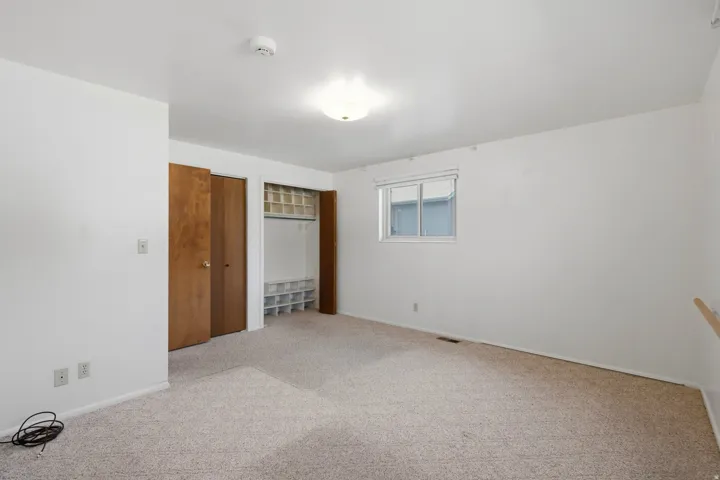Spacious bedroom featuring neutral carpeting, two wood-finish closet doors with shelving, a large window with blinds, and multiple electrical outlets