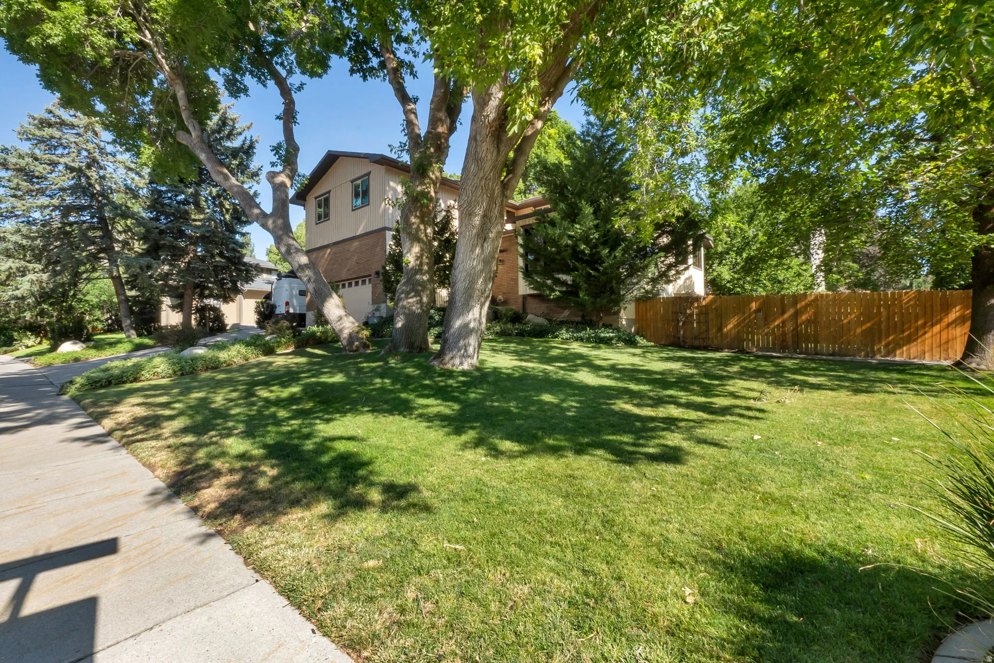 Expansive front lawn with mature shade trees, showcasing a two-story residence with a brick and stucco facade, a two-car garage, and a wood fence enclosing the side yard