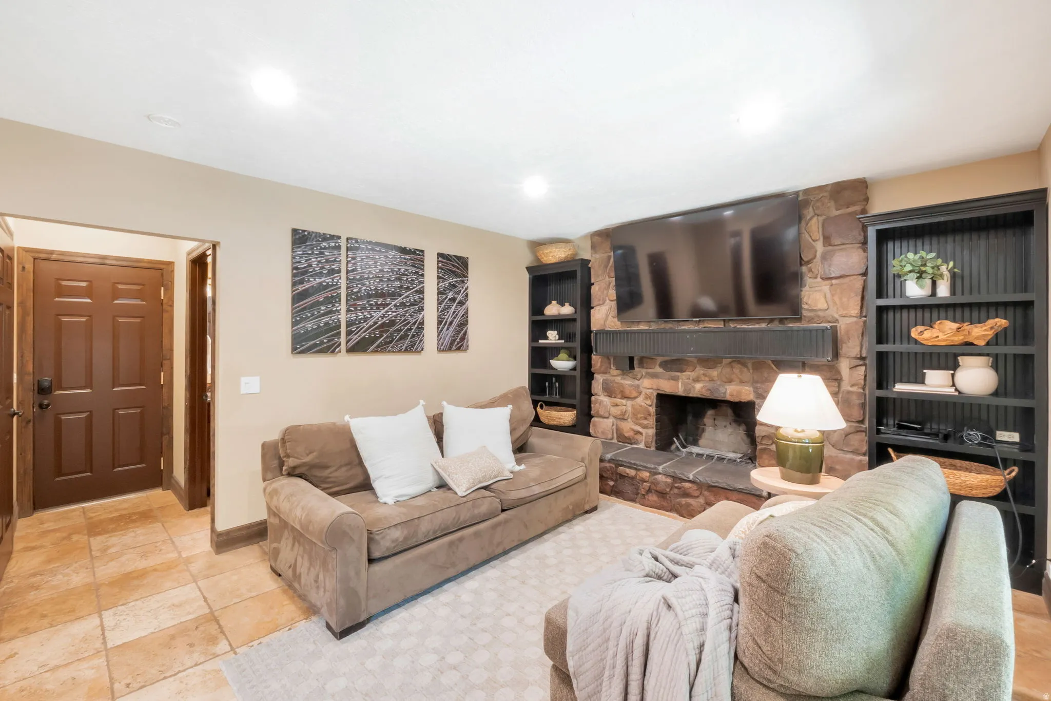 Living area featuring a stone-clad fireplace with a dark wood mantel, recessed lighting, built-in shelving, tile flooring, and a paneled entry door