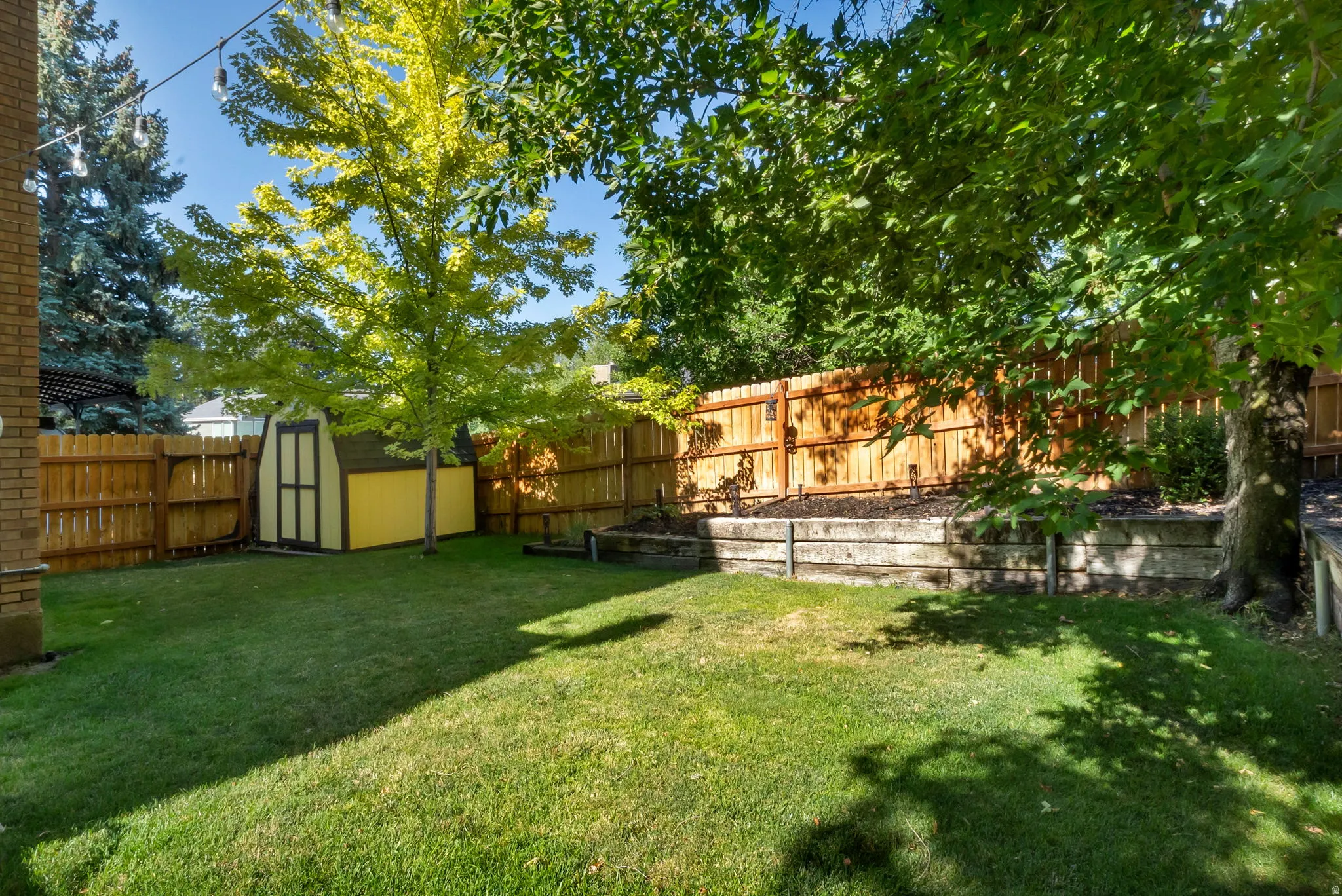 Spacious rear yard featuring mature trees, a wooden privacy fence, a yellow storage shed, and a tiered retaining wall