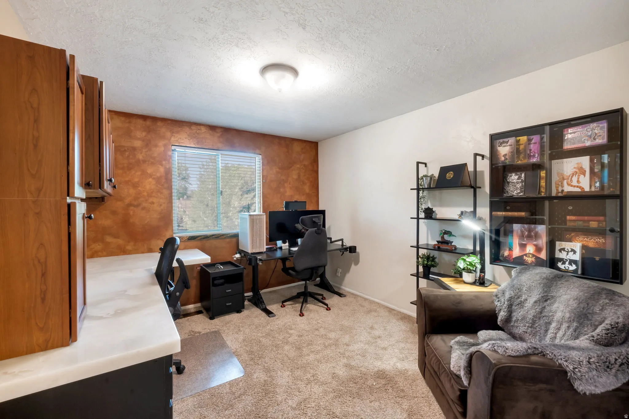 Room featuring textured accent wall, window with horizontal blinds, built-in cabinetry with wood-finish doors, light carpet, and a flush-mount ceiling light fixture