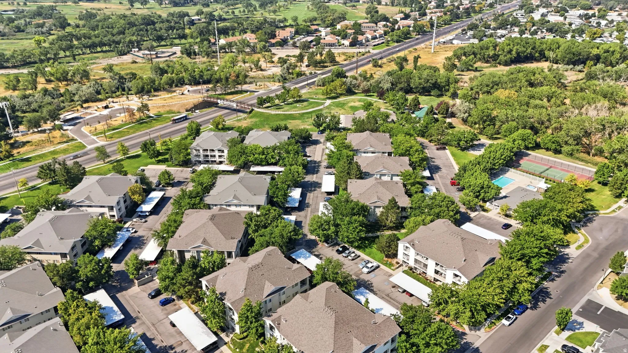 Aerial overview of a multi-building residential complex featuring multiple parking areas, mature landscaping, and a nearby recreational area with a swimming pool and tennis courts