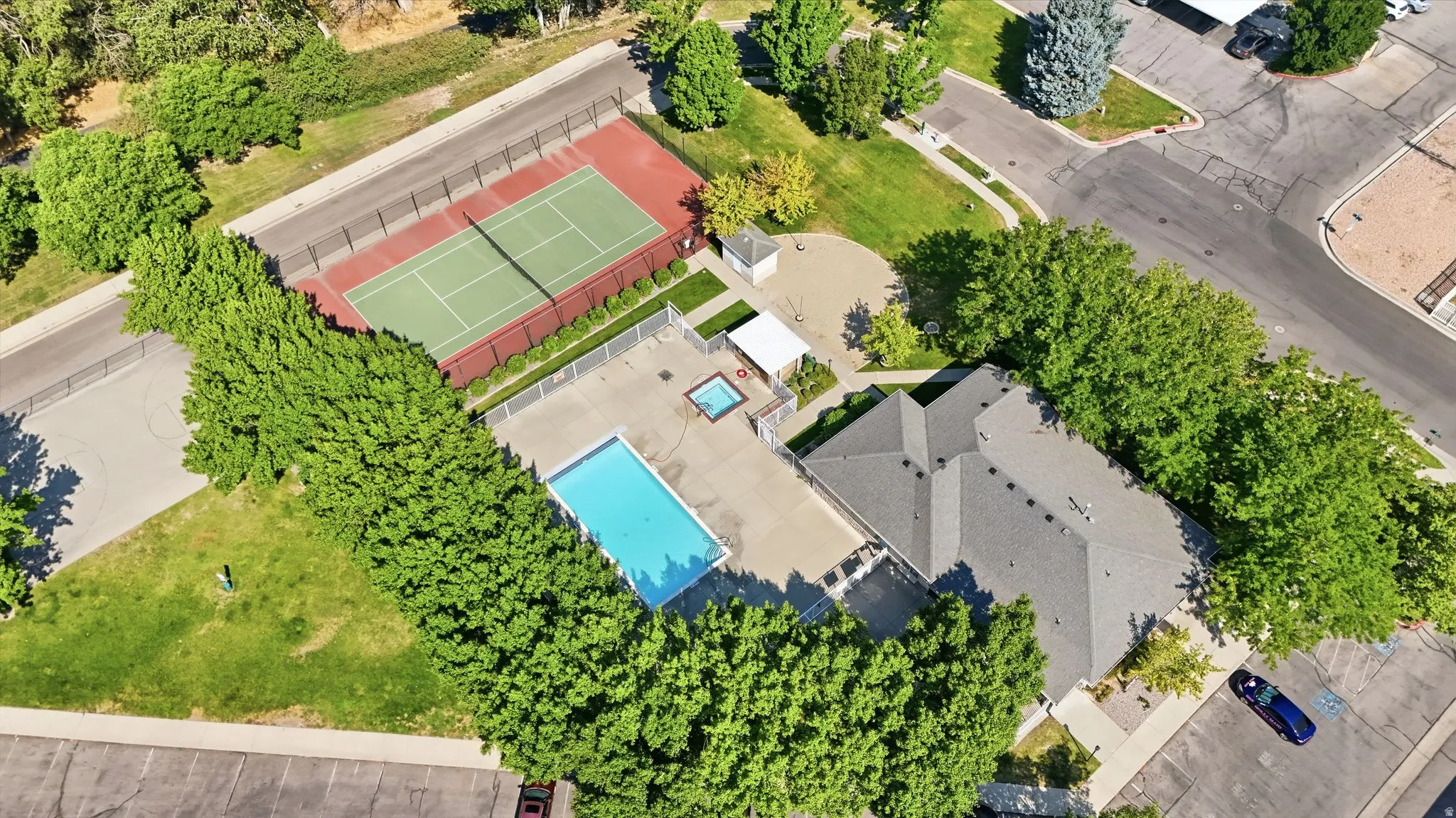 Aerial view showcasing resort-style amenities including a sparkling swimming pool, professional-grade tennis courts with red clay surface, manicured lawn with mature tree landscaping, and a modern clubhouse structure, all nestled within lush grounds and a