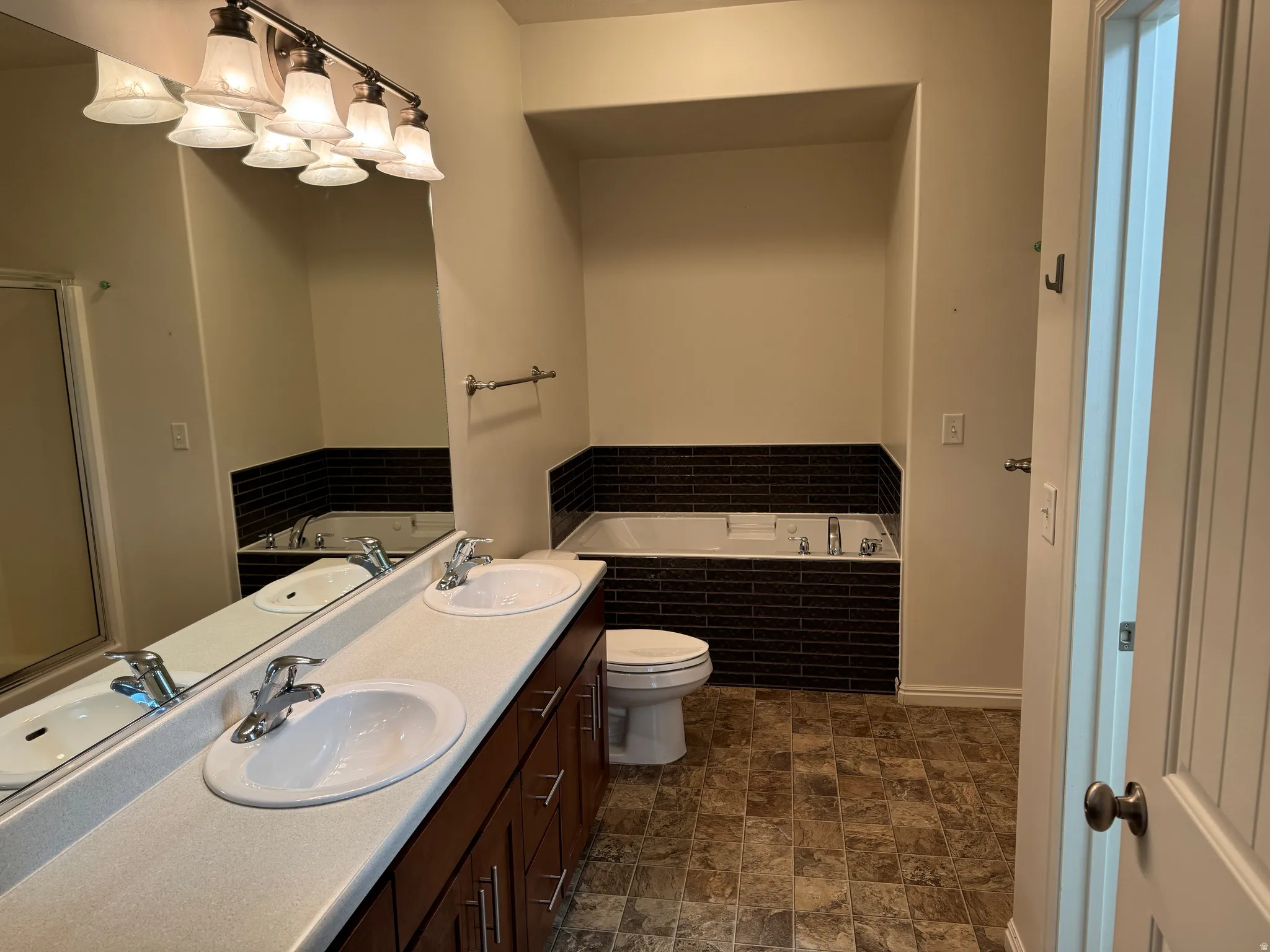 Bathroom featuring a dual vanity with undermount sinks, a built-in soaking tub with dark tile surround, a toilet, and patterned flooring