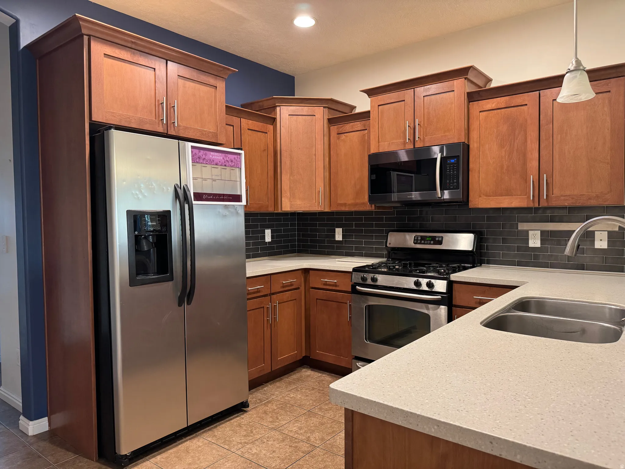 Kitchen featuring wood-finish cabinetry, stainless steel appliances, solid surface countertops, a dark subway tile backsplash, and tile flooring