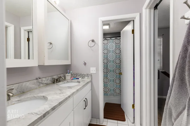 Spa-inspired primary bathroom featuring a marble-topped double vanity with dual sinks and chrome fixtures, large framed mirrors, white cabinetry, geometric tile-accented shower enclosure with glass doors, and wood-finish flooring throughout