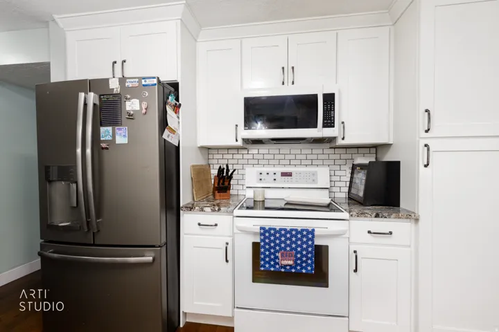 Kitchen featuring white shaker-style cabinetry with dark hardware, a stainless steel refrigerator, white electric range with overhead microwave, white subway tile backsplash, and granite-style countertops