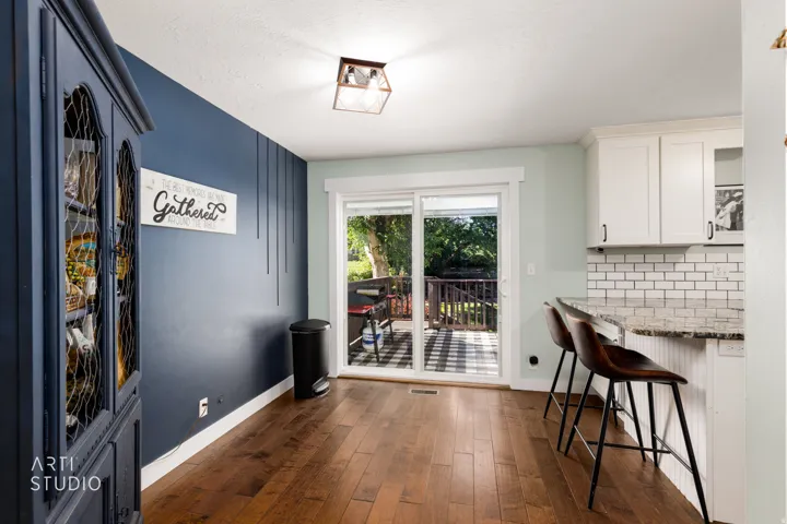 Wood-finish flooring throughout, featuring a kitchen bar with seating, white shaker cabinetry, subway tile backsplash, and sliding glass door access to an outdoor deck