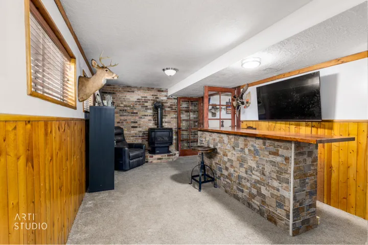Finished basement recreation area featuring exposed brick accent wall, wood-finish wainscoting, built-in shelving with display cabinetry, wet bar with counter seating, recessed ceiling lighting, and carpeted flooring throughout